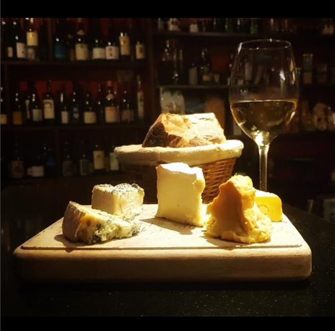 Planche de fromages affinés et panier de pain maison devant les rayonnages de bouteilles, avec un verre de vin blanc